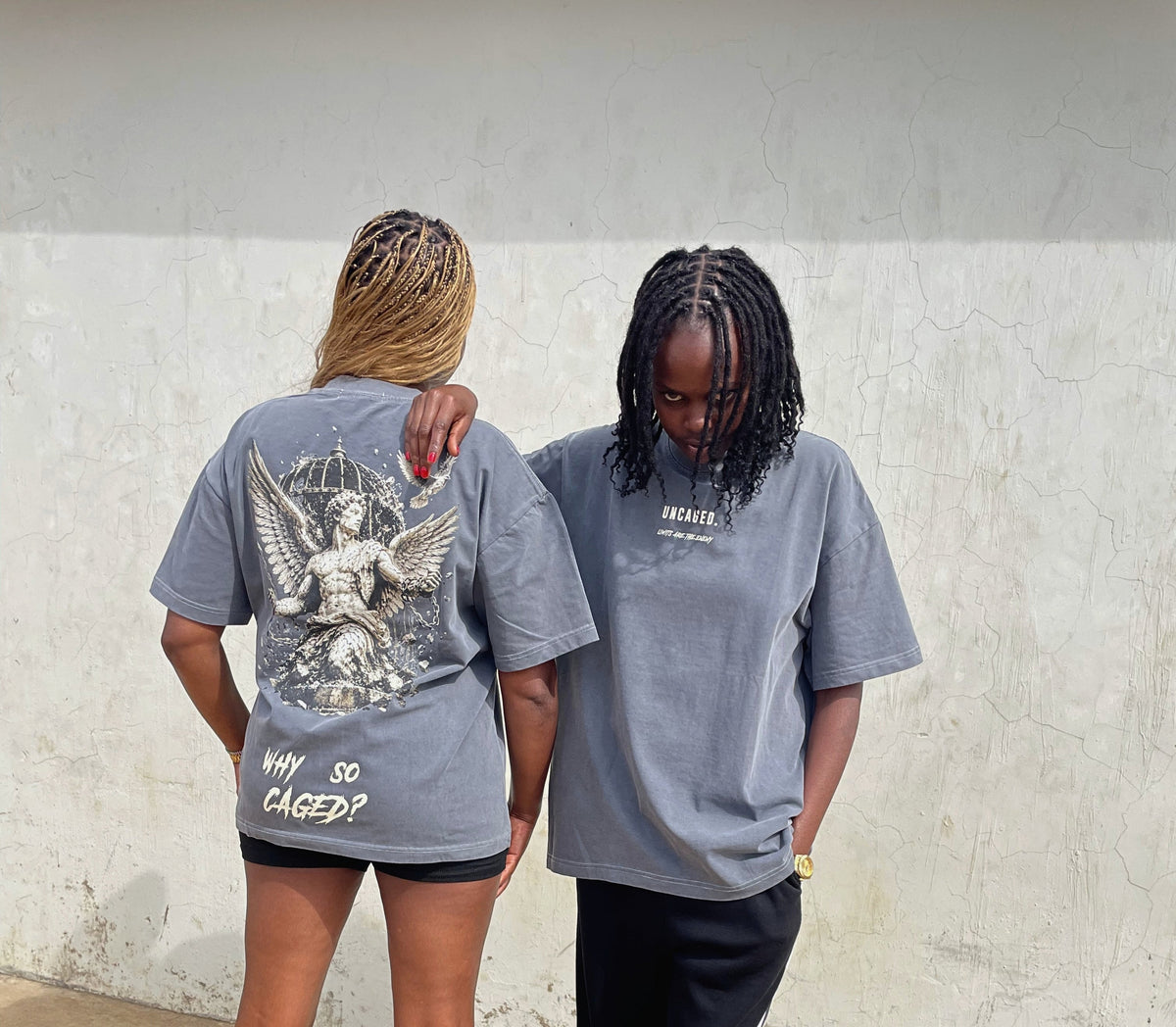 Two models wearing oversized gray graphic t-shirts in an outdoor setting against a textured concrete wall. The model on the left shows the back of the t-shirt featuring a large white angel graphic and the text 'WHY SO CAGED?'.